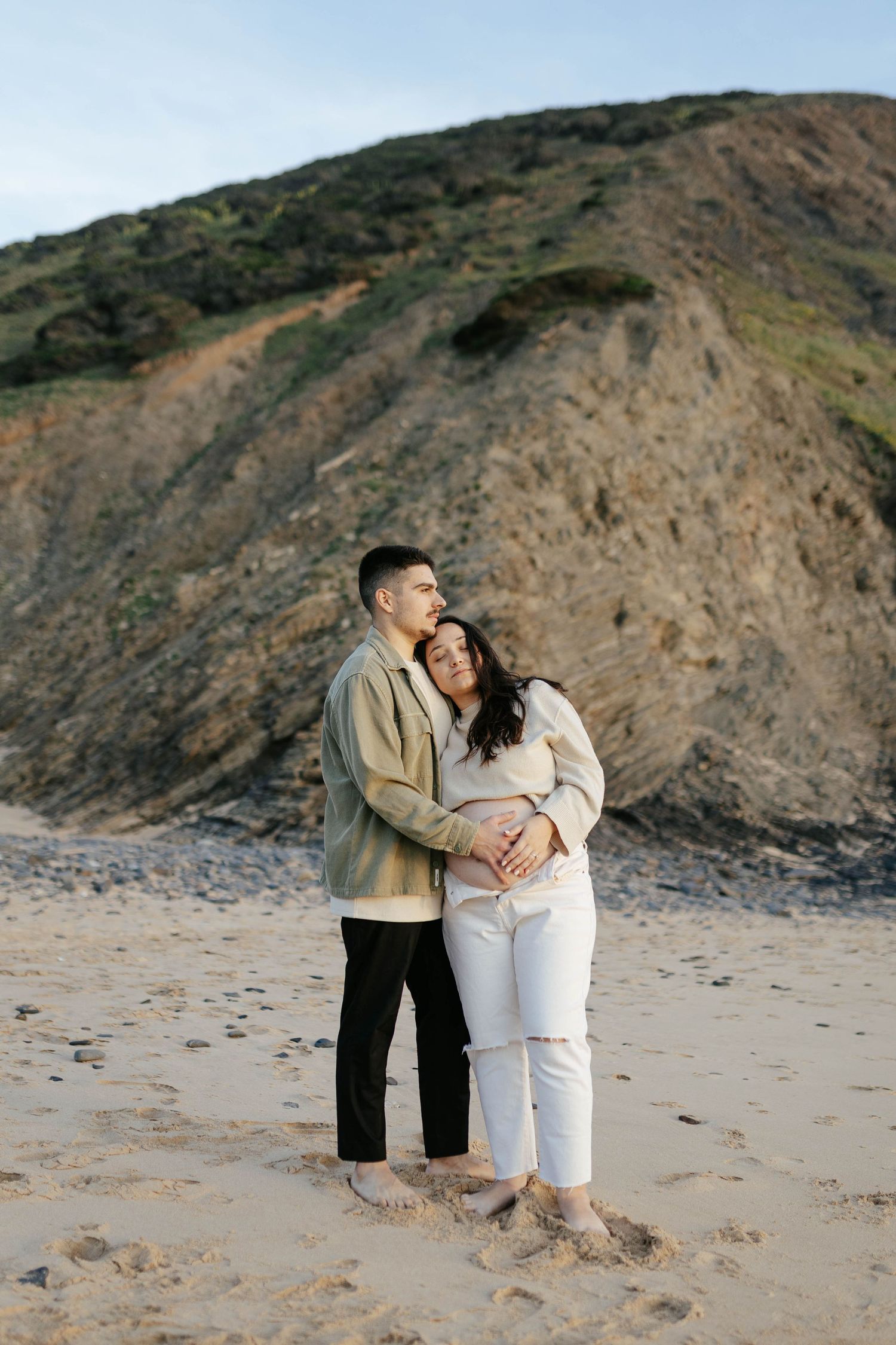 A couple shares an intimate embrace on a sandy beach with dramatic coastal cliffs rising in the background.