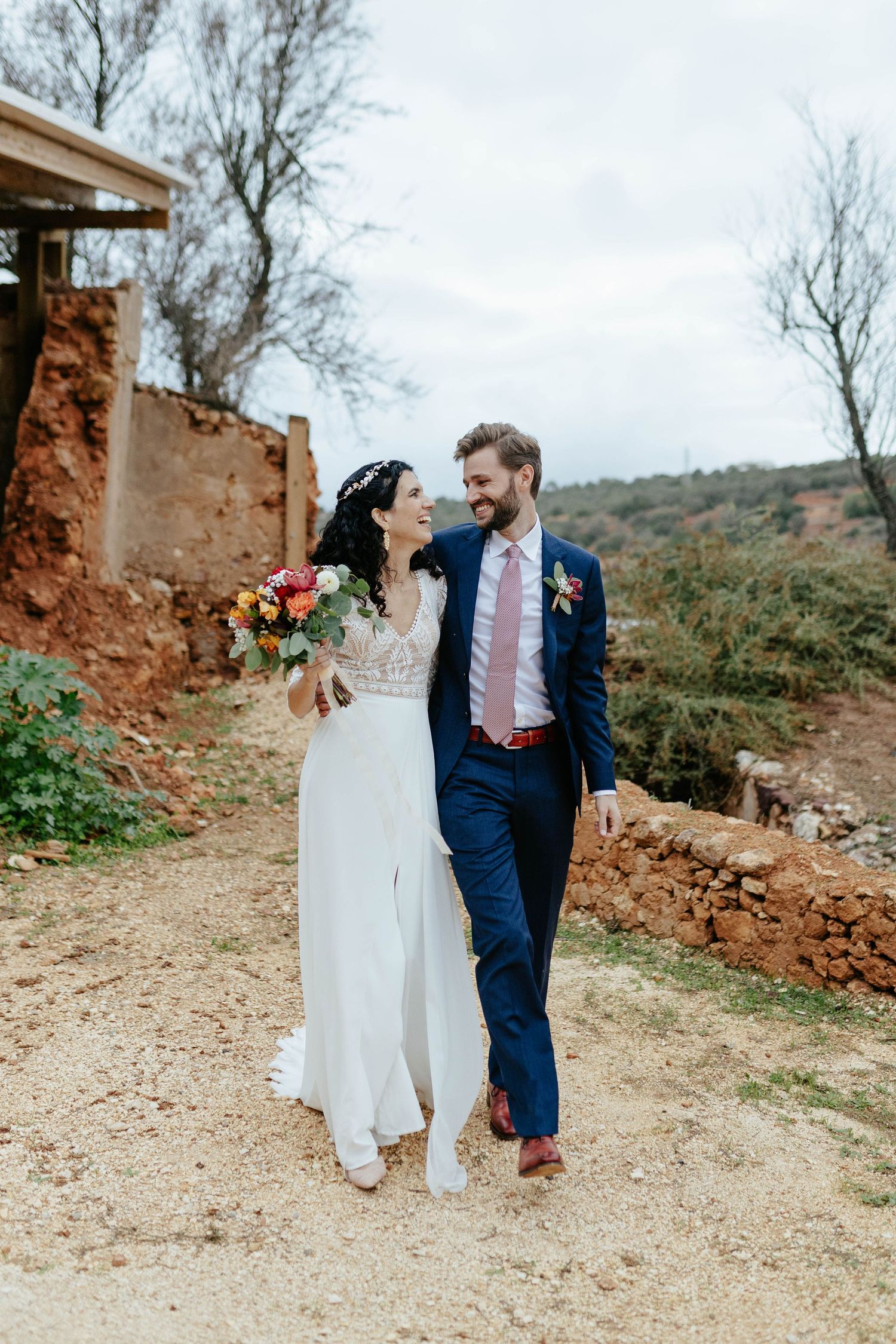 A couple walking together on a dirt path in formal attire with orange cliffs and bare trees in the background.