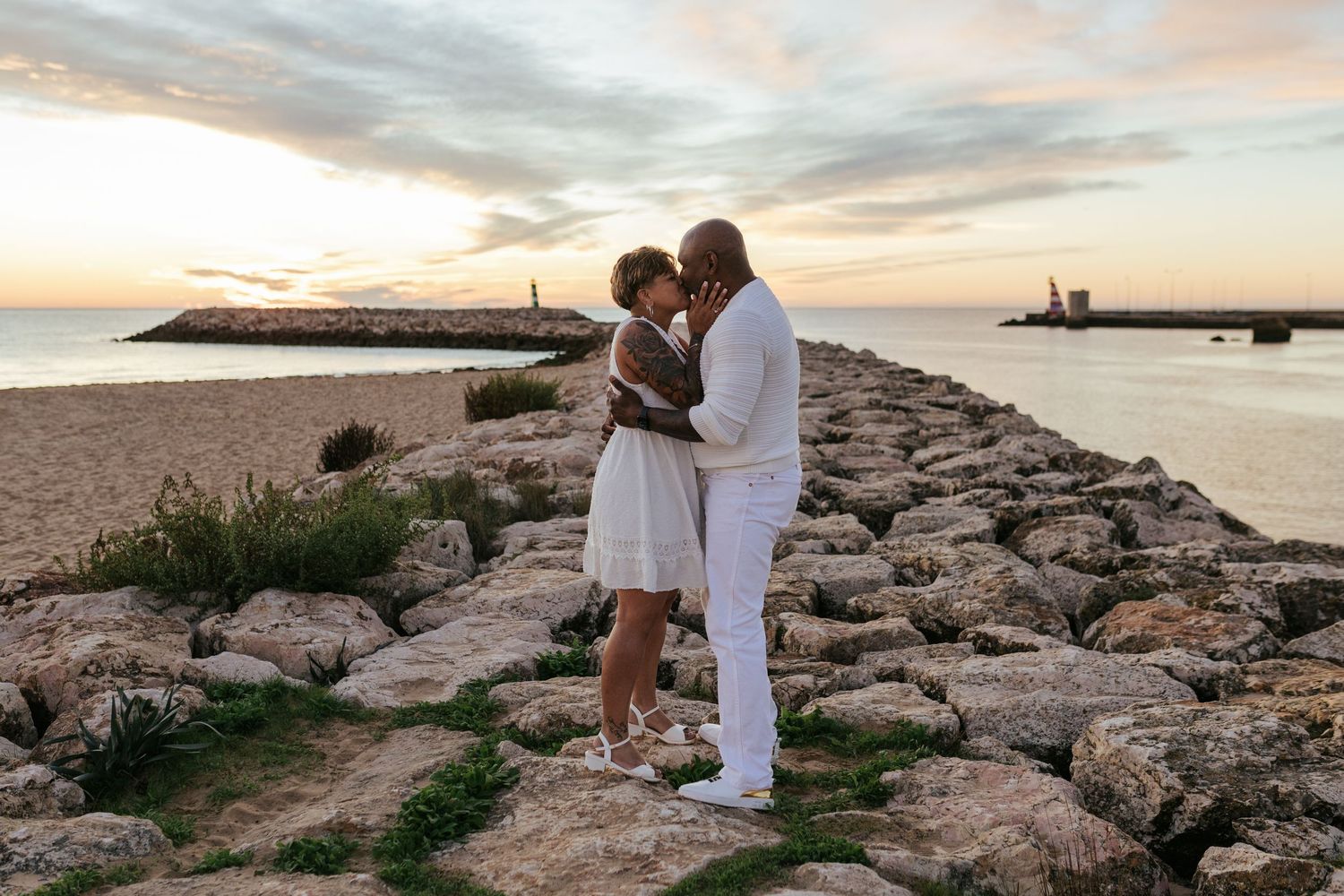 Romantic couple embracing on rocky jetty at sunrise with lighthouse in background. Meia Praia, Lagos, Algarve, Portugal.
