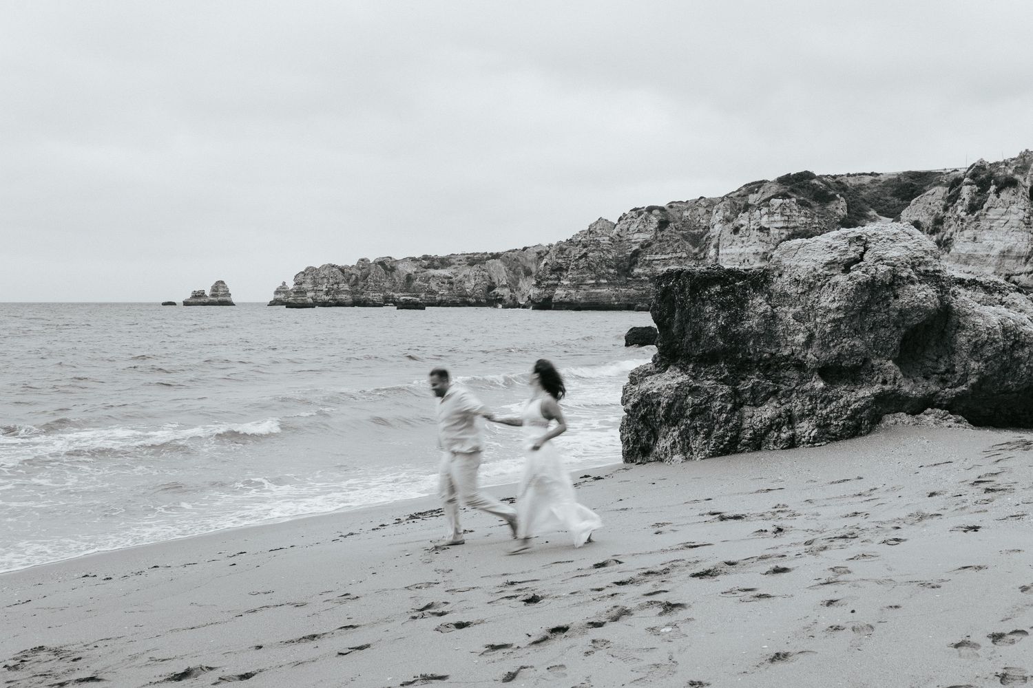 Black and white photos of a couple walking and playing on a rocky beach during their engagement photoshoot.