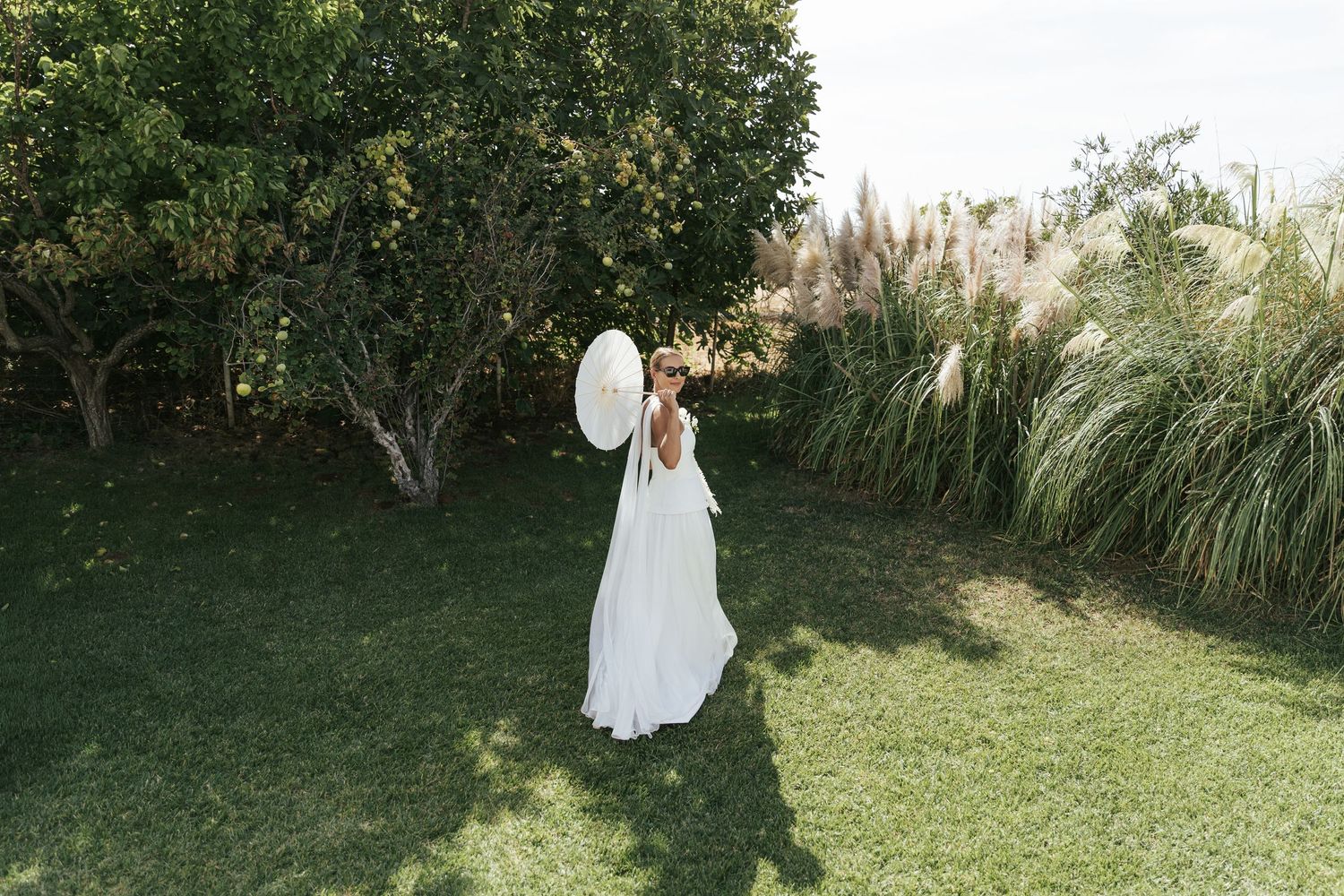 Bride in white gown holding parasol umbrella in lush green garden.