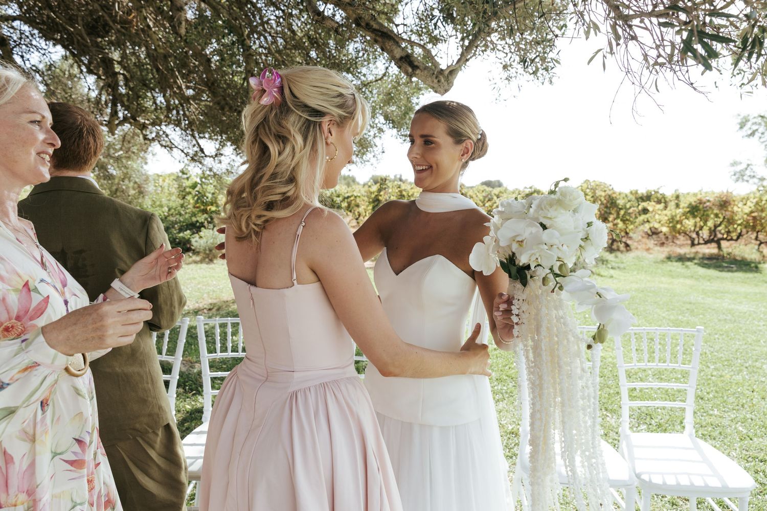 Bride in white gown holding bouquet chats with bridesmaid in pink dress at outdoor garden wedding ceremony.