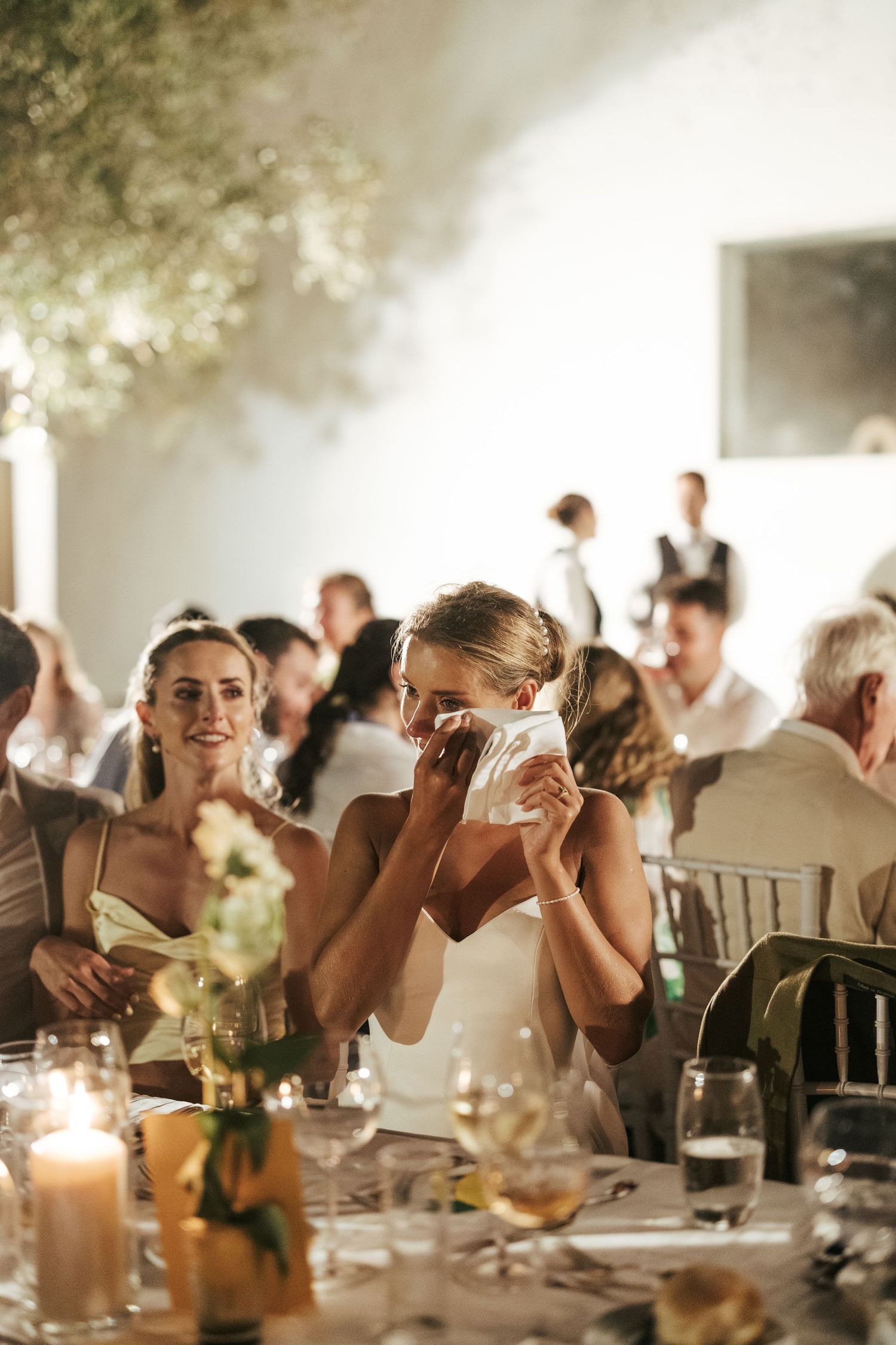 Emotional bride dabbing tears with napkin at wedding reception table surrounded by candles and floral centerpieces.