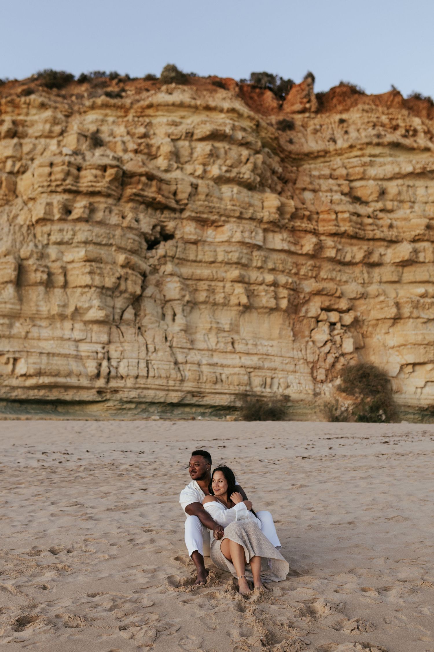 A couple sits close together on sandy beach with dramatic layered cliff face rising behind them. Praia do Porto de Mós, Lagos, Algarve, Port