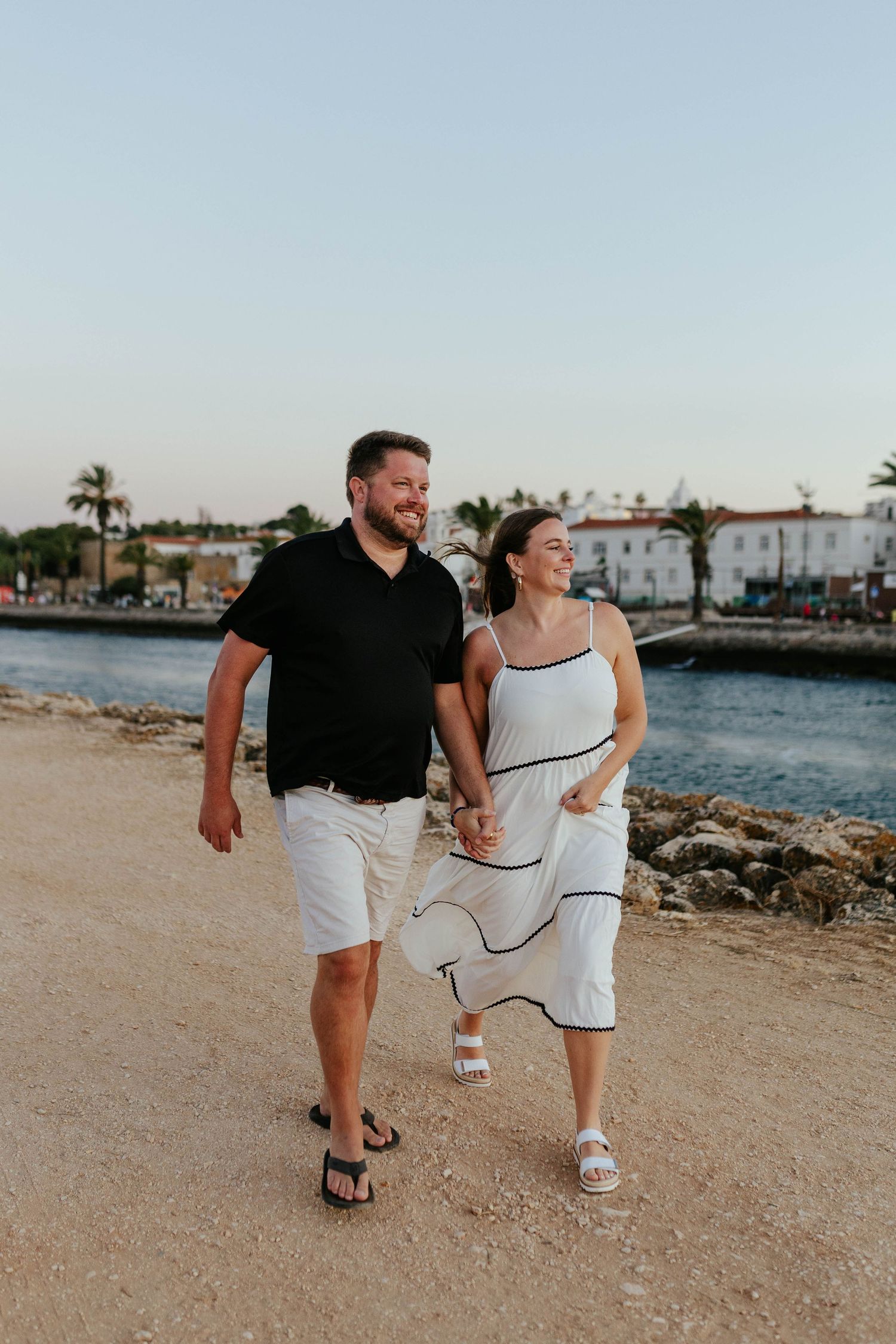 Couple strolling along waterfront path with palm trees and buildings in background. Meia Praia, Lagos, Algarve, Portugal.