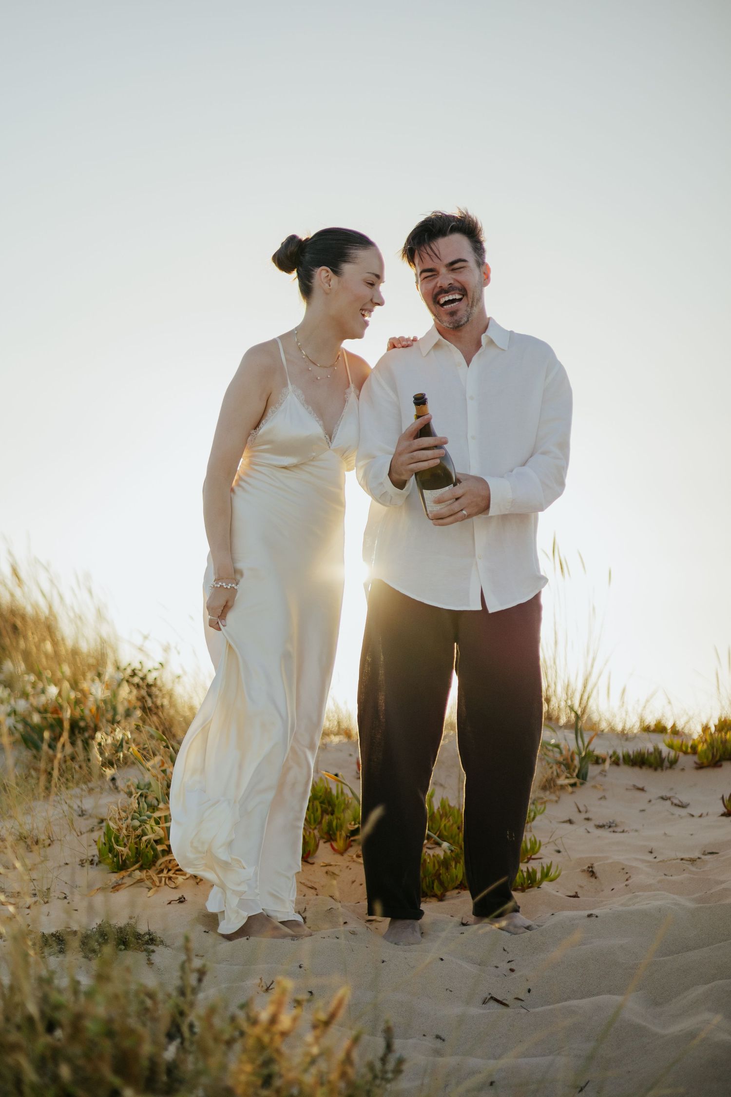 Joyful couple laughing together on sandy beach at sunset while holding a bottle. Meia Praia, Lagos, Algarve, Portugal.