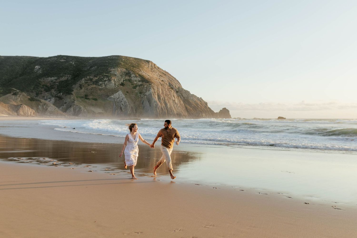 Two people walk along a misty beach with dramatic coastal cliffs and crashing waves in the background at sunset.
