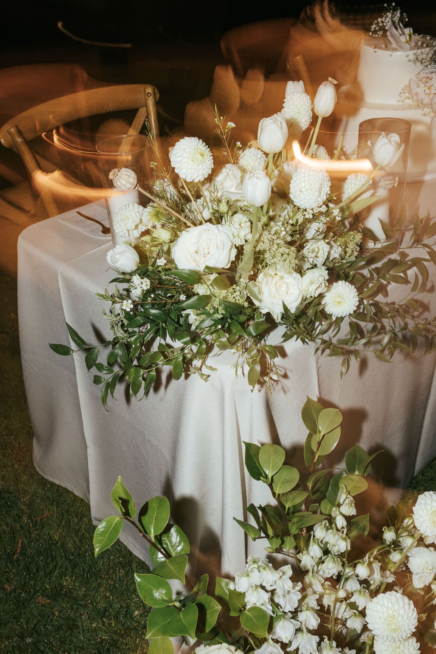 Elegant white floral centerpiece with roses and greenery on table at evening wedding reception.