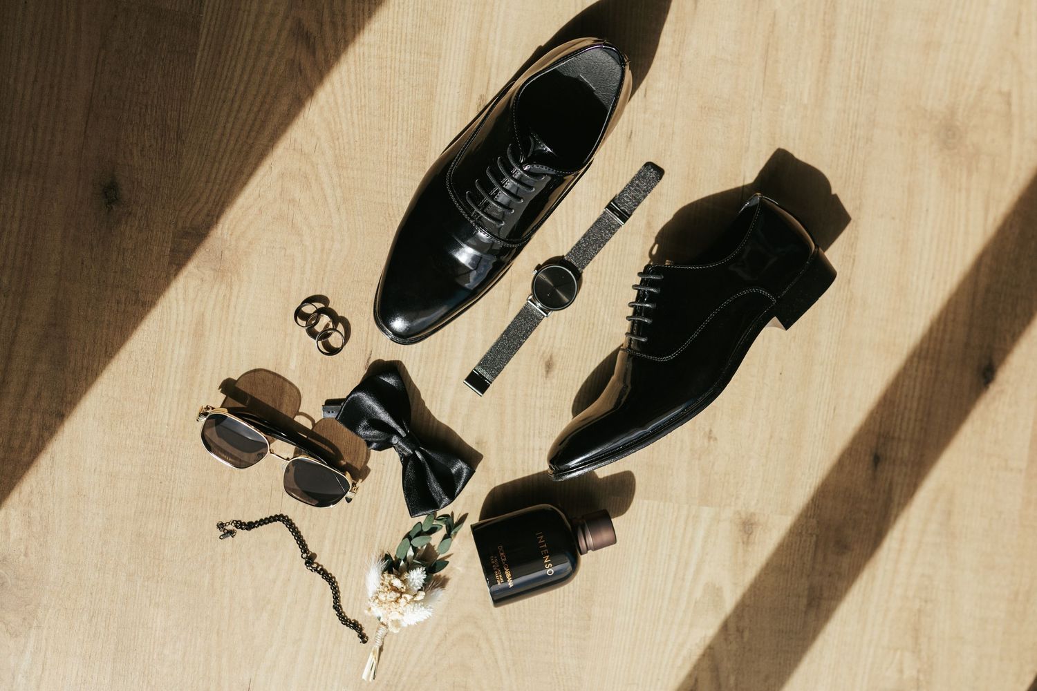 Flat lay of formal black dress shoes with accessories including watch, sunglasses, and tie on wooden floor.