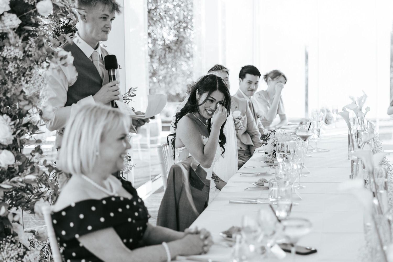 Black and white wedding reception photo of guests laughing during a heartfelt speech at an elegant dining table.