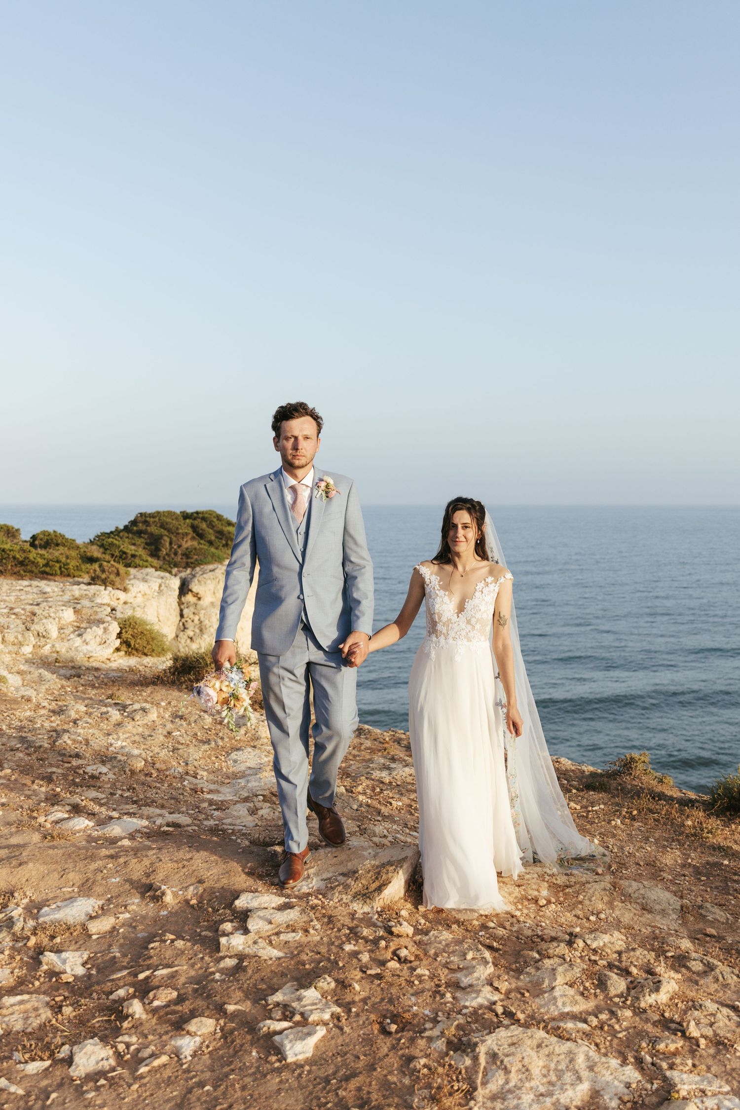 Bride and groom holding hands on rocky coastal cliff, wearing white gown and light blue suit, ocean backdrop.