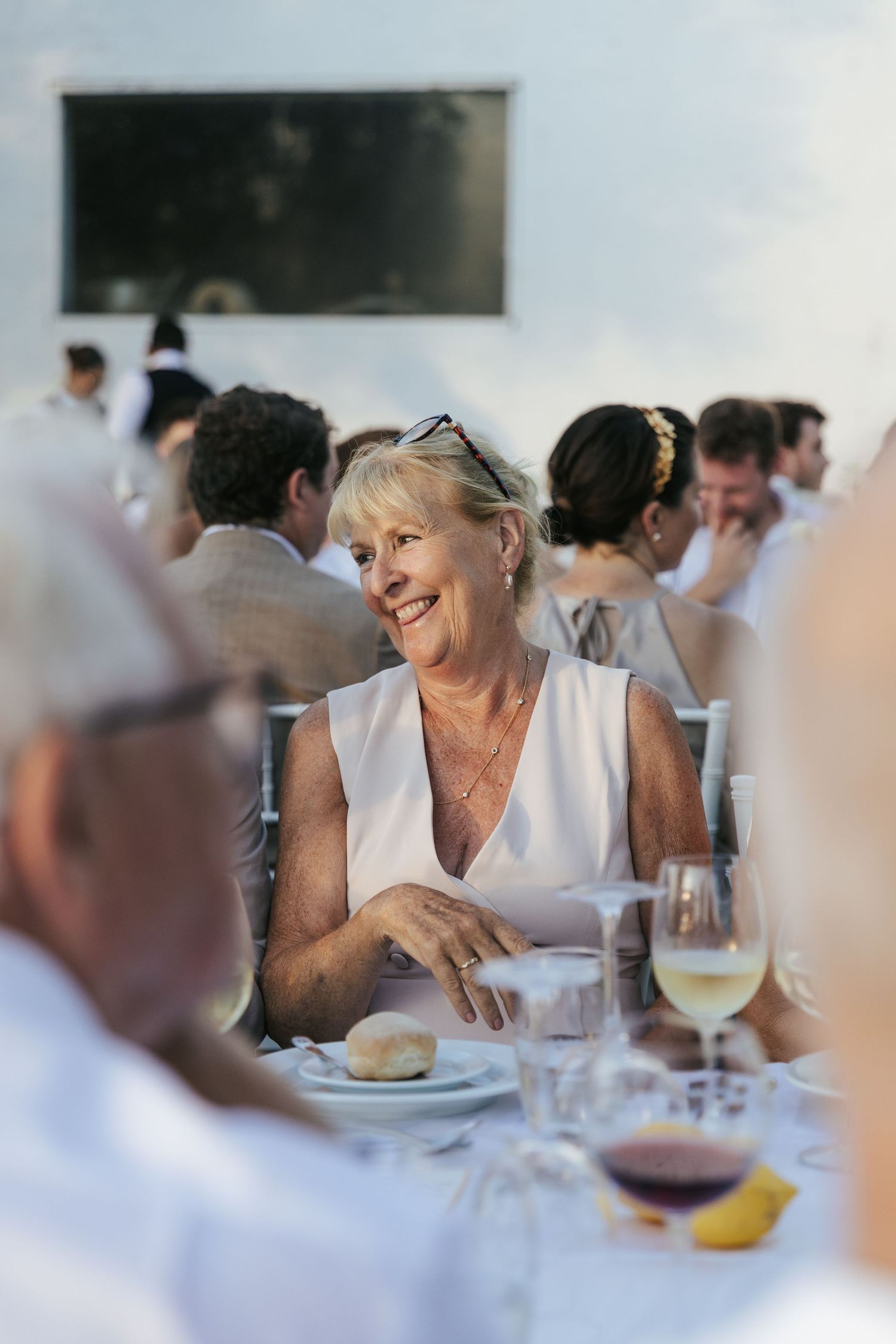 Smiling older woman in white dress at outdoor wedding reception dinner table with wine glasses.