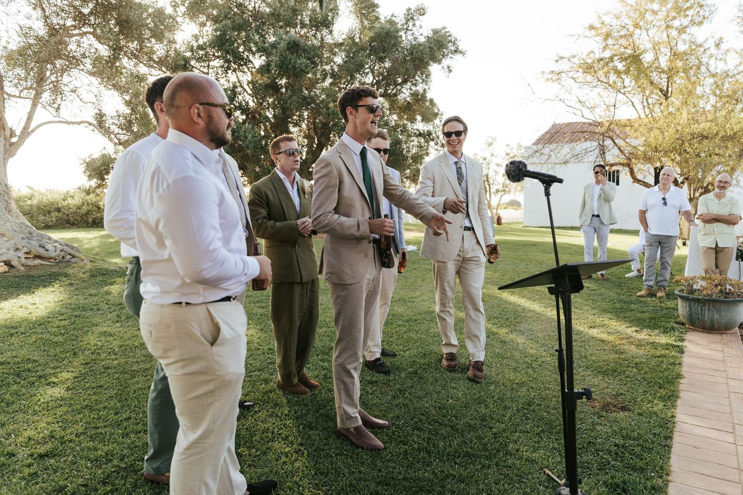Groomsmen in tan and olive suits gathered on lawn during outdoor wedding ceremony.