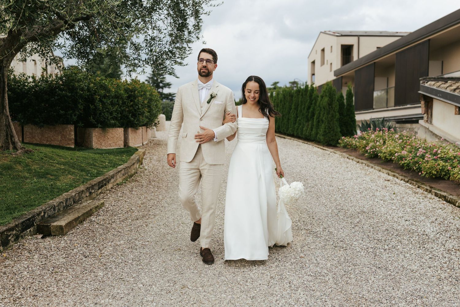 Newlyweds walking together on gravel path surrounded by lush garden and architecture.