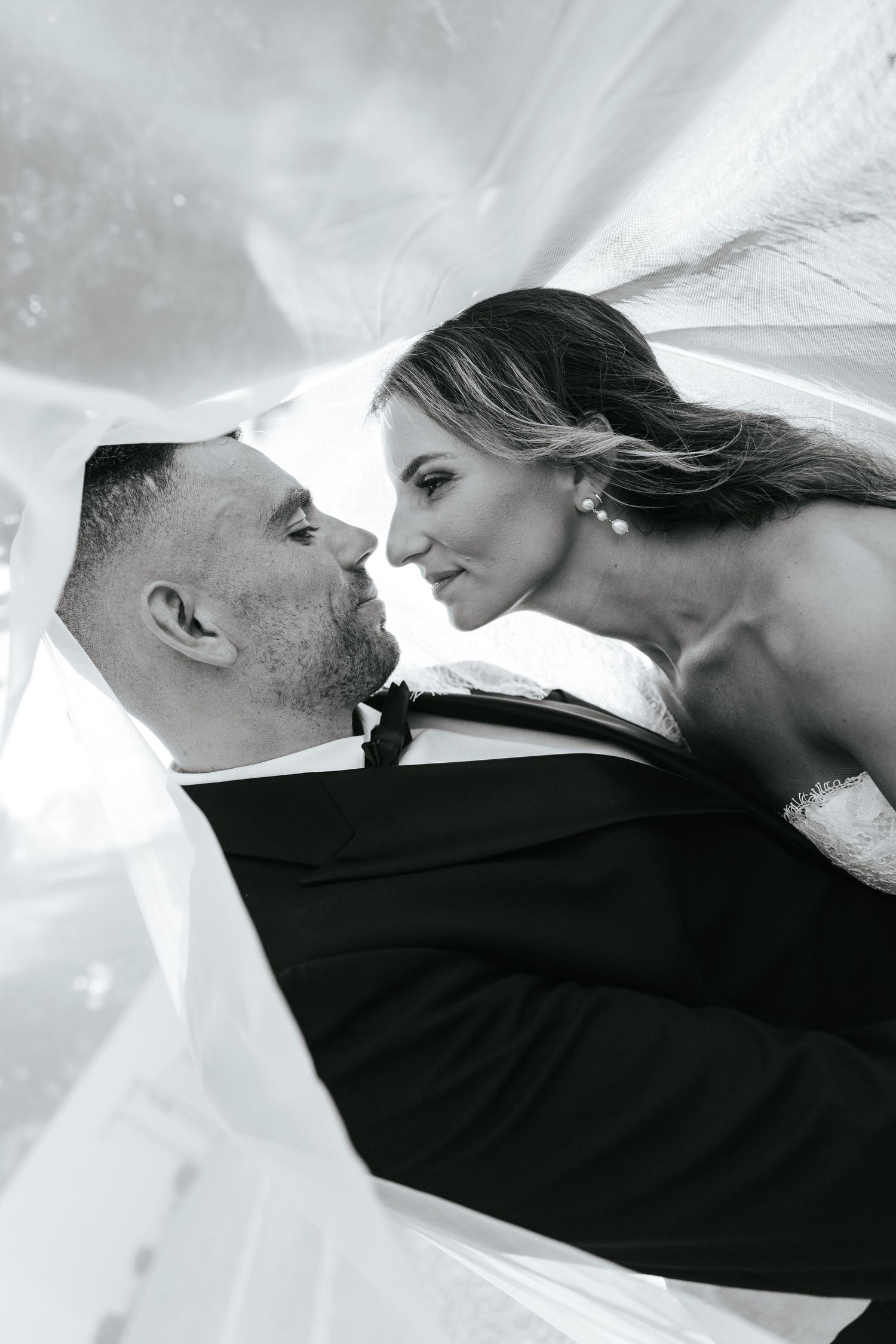 Black and white wedding photo of bride and groom almost kissing under flowing veil.
