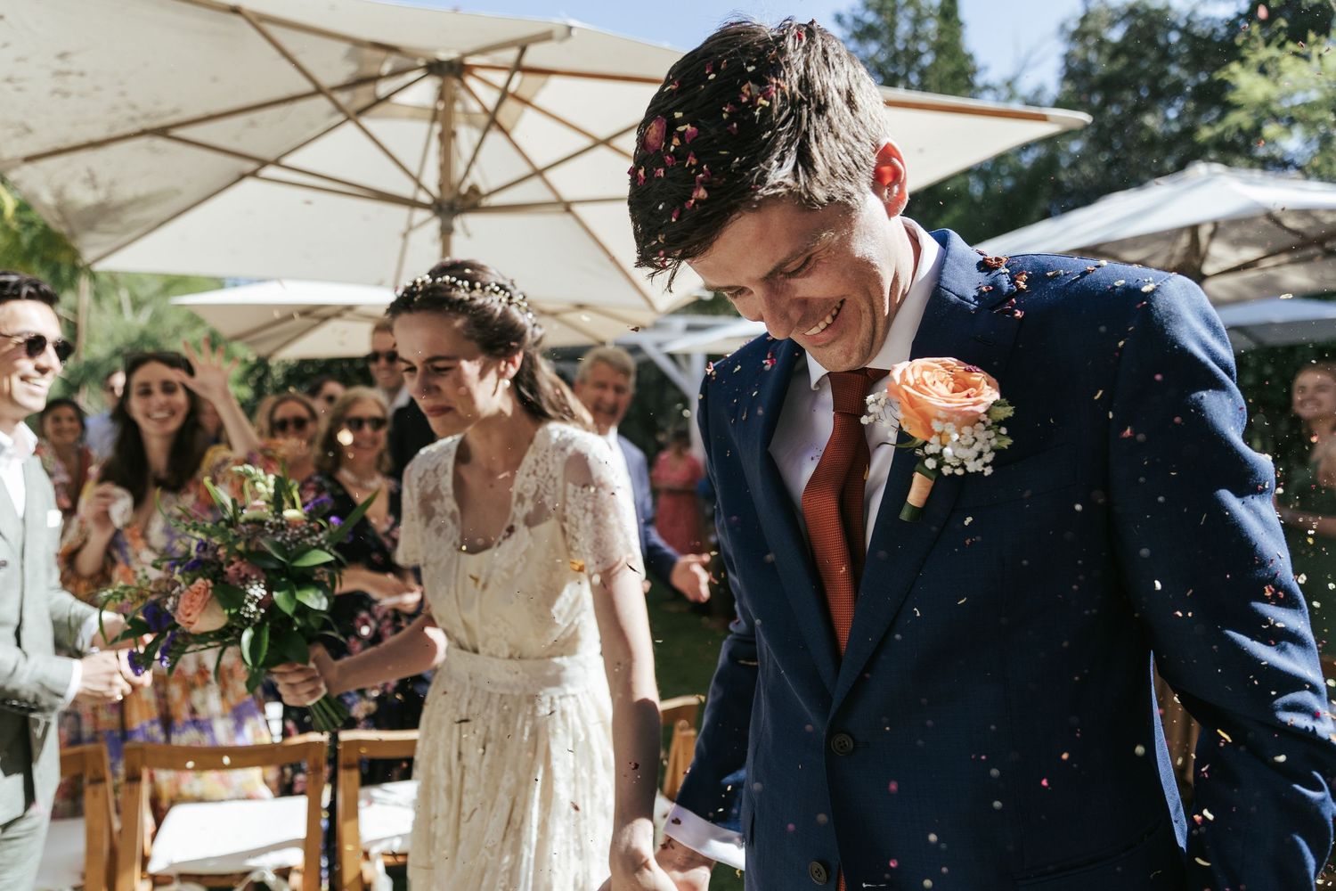 Newlyweds walk through a joyful shower of confetti at an outdoor wedding reception with white umbrellas in background.