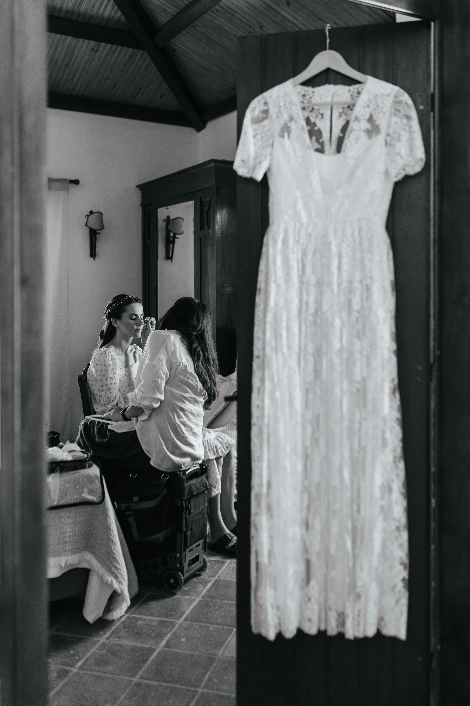 Black and white photo of a white lace wedding dress hanging while preparations take place in background.