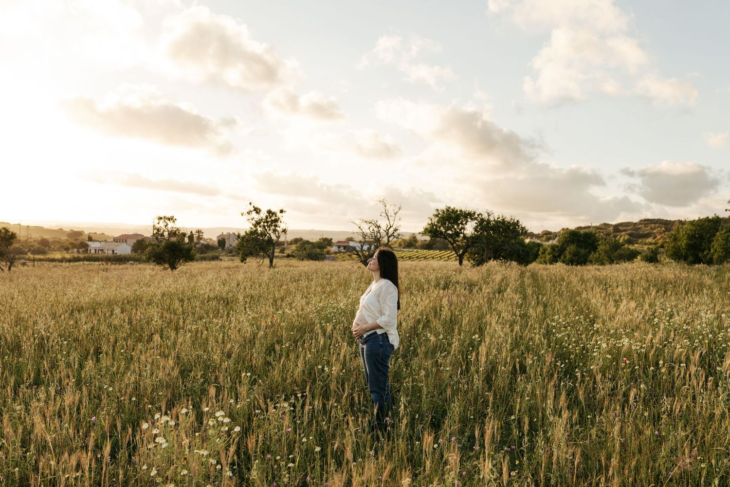 A person stands in a golden field at sunset with scattered trees and rolling hills in the background.