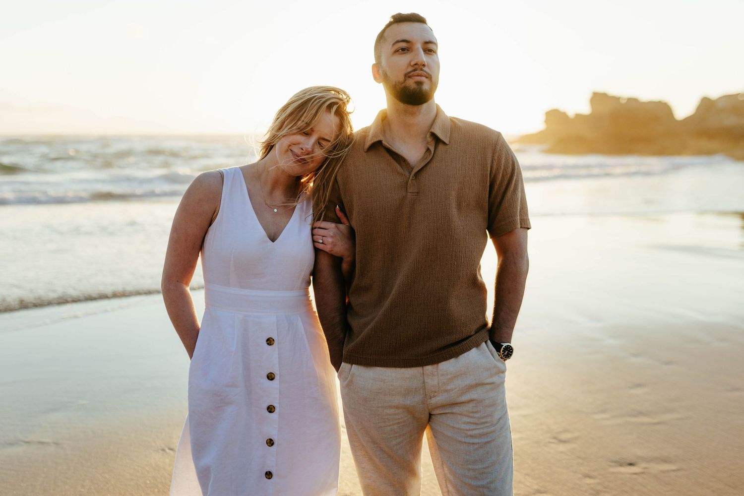 A couple enjoys a romantic moment at sunset on a sandy beach with ocean waves and rocks in the background.