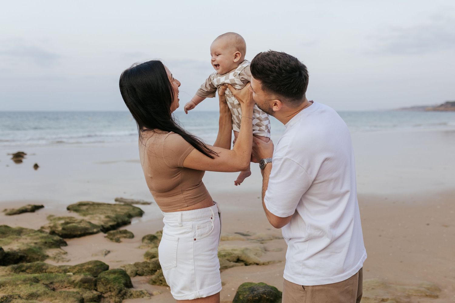 Family enjoying a tender moment together at a rocky beach with ocean waves and cloudy sky in the background.