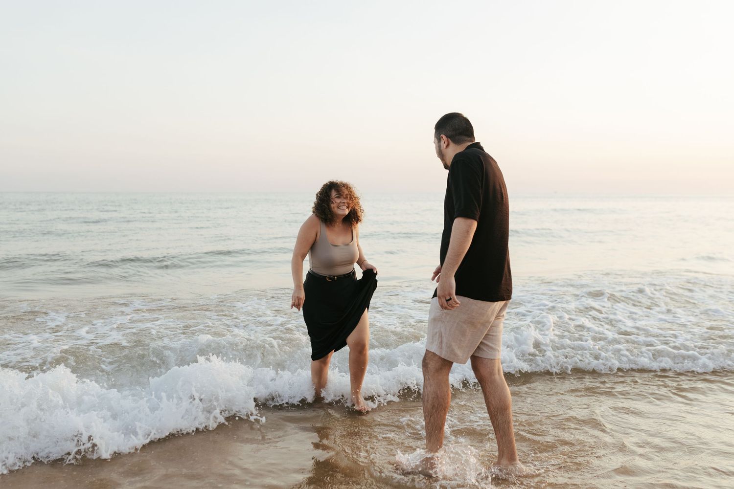 A couple walks together at the beach during sunset while ocean waves crash against the shore.