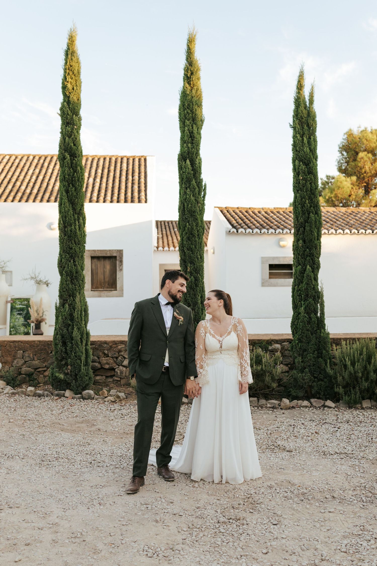 A couple stands together on a gravel path beside tall cypress trees and white stucco buildings with terracotta roofs.