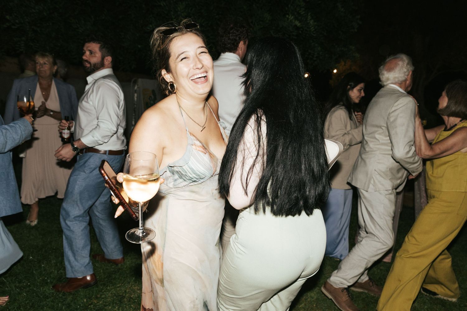 Two women laughing and dancing together at an outdoor evening party, one holding a cocktail glass.