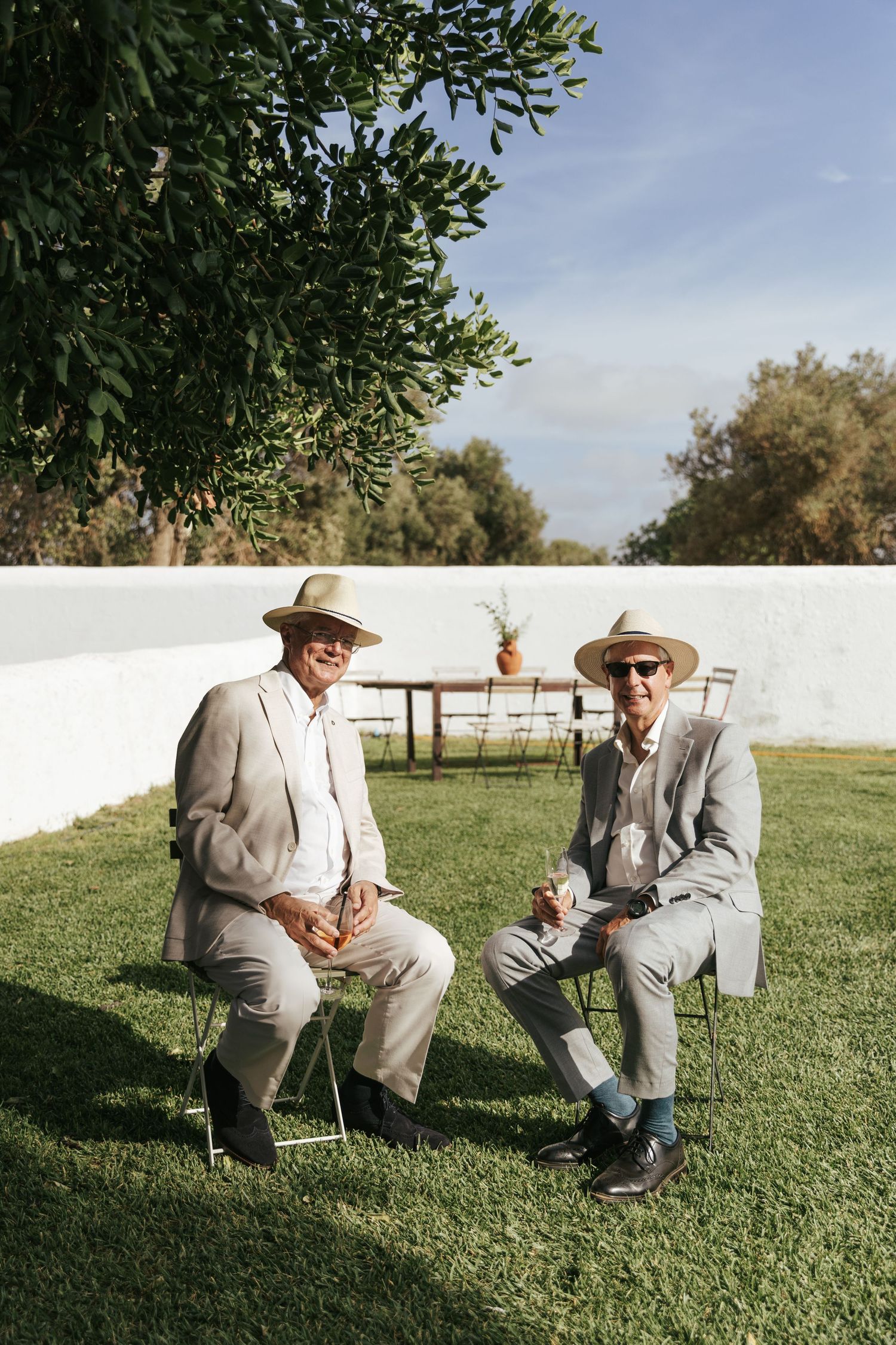 Two elderly men in linen suits and straw hats sit on chairs in a sunny garden with white walls.