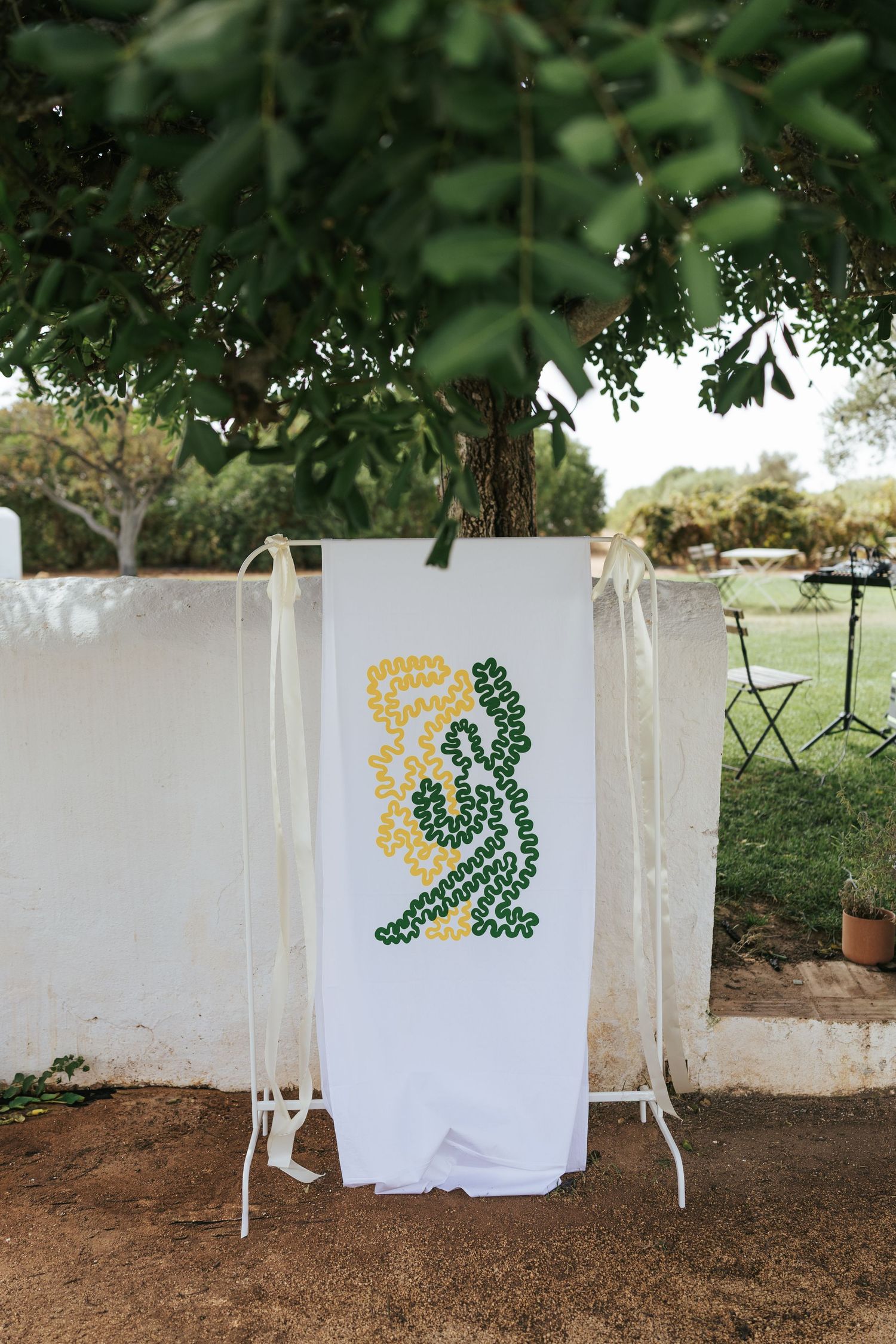 White banner with green and yellow floral design displayed outdoors on a stand near a tree at a garden event.