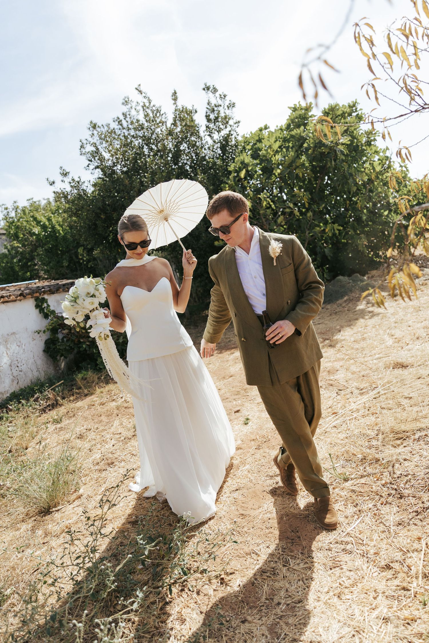 Bride and groom walking outdoors in sunglasses, she holding white parasol and bouquet in strapless gown, he in brown suit.