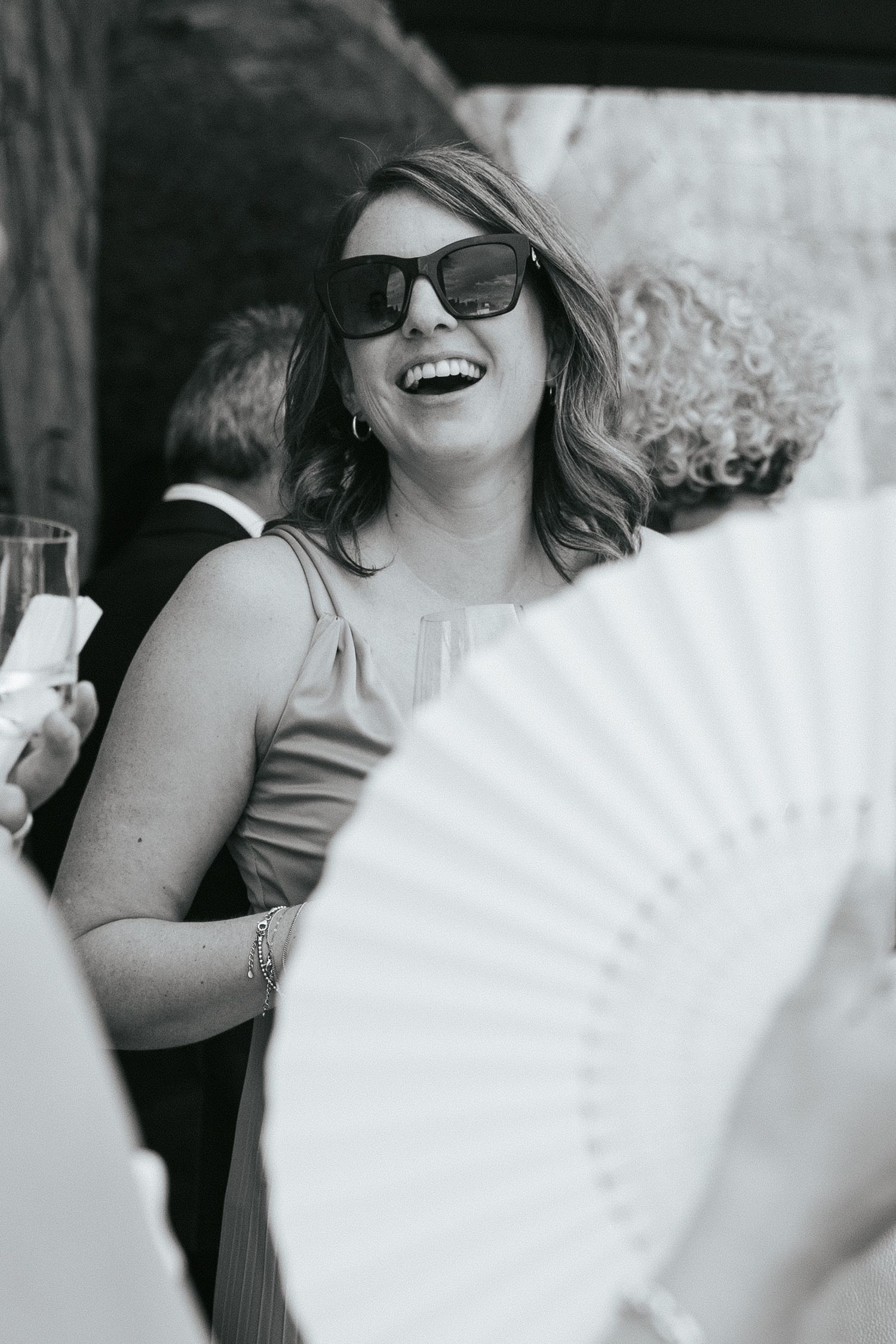 Guest with sunglasses smiling while holding white decorative fan at outdoor celebration.