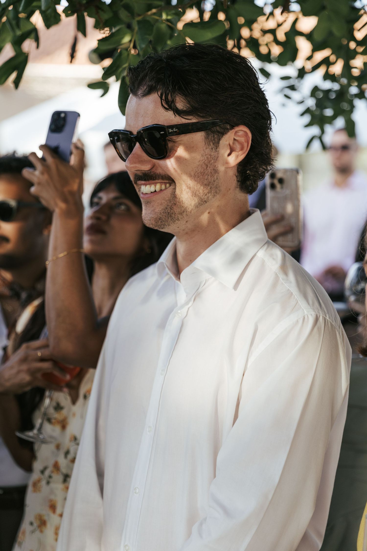 Man in white shirt and sunglasses smiling at outdoor event with people in background.