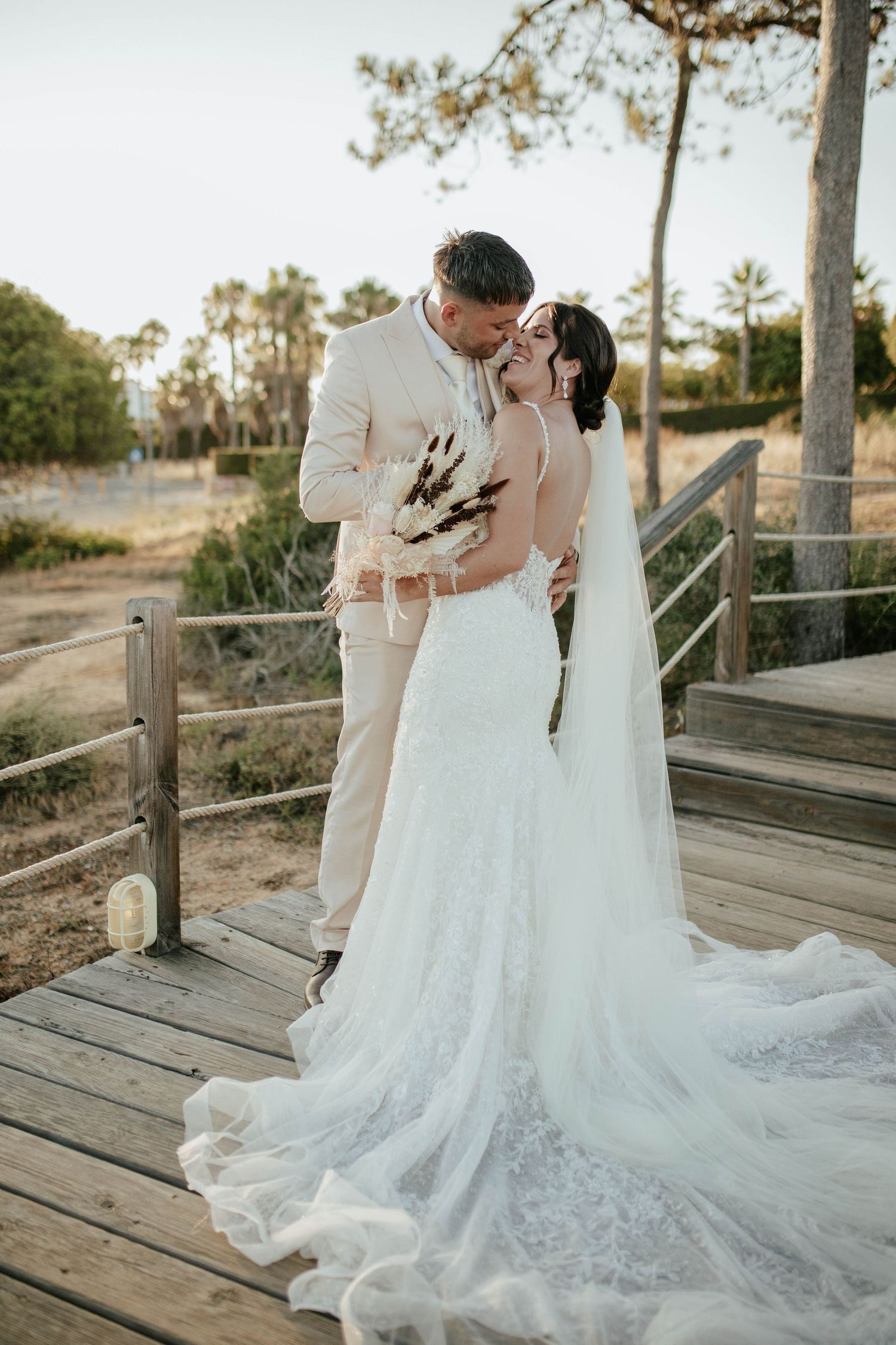 A romantic wedding couple embraces on a wooden boardwalk with palm trees and golden sunlight in the background.