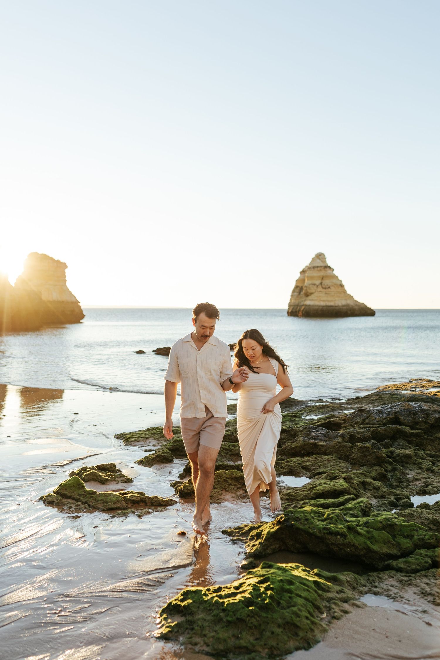 Couple walking together on mossy rocks at a scenic beach with coastal rock formations during golden hour sunset.