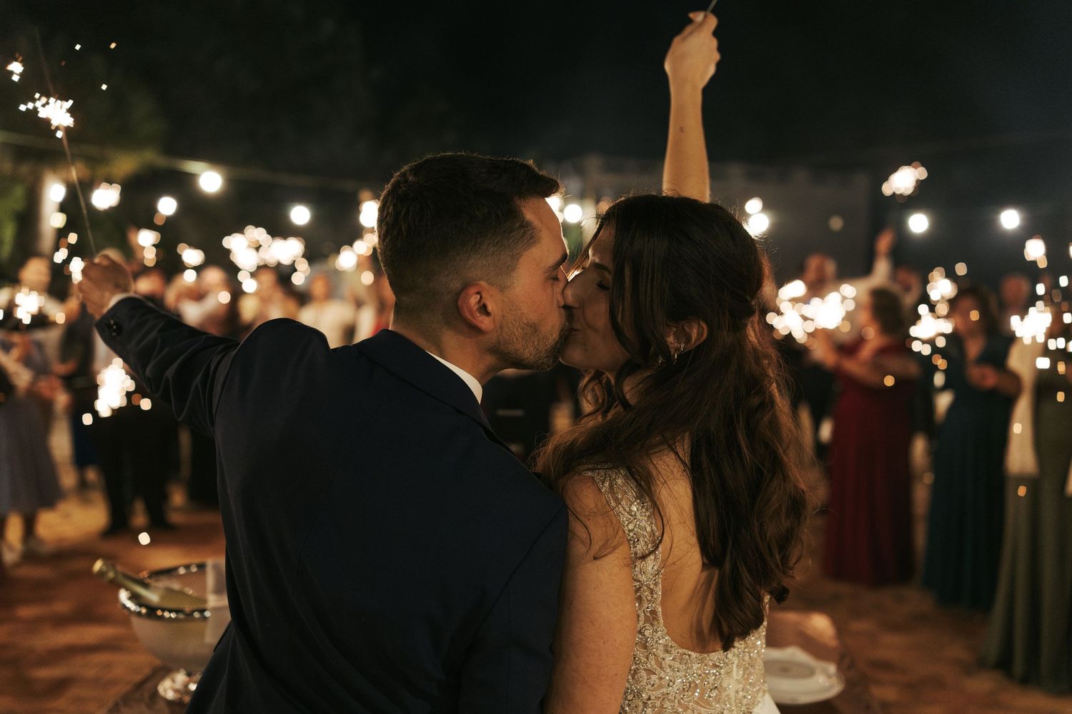 Newlyweds share an intimate moment surrounded by sparklers and wedding guests during their evening celebration.