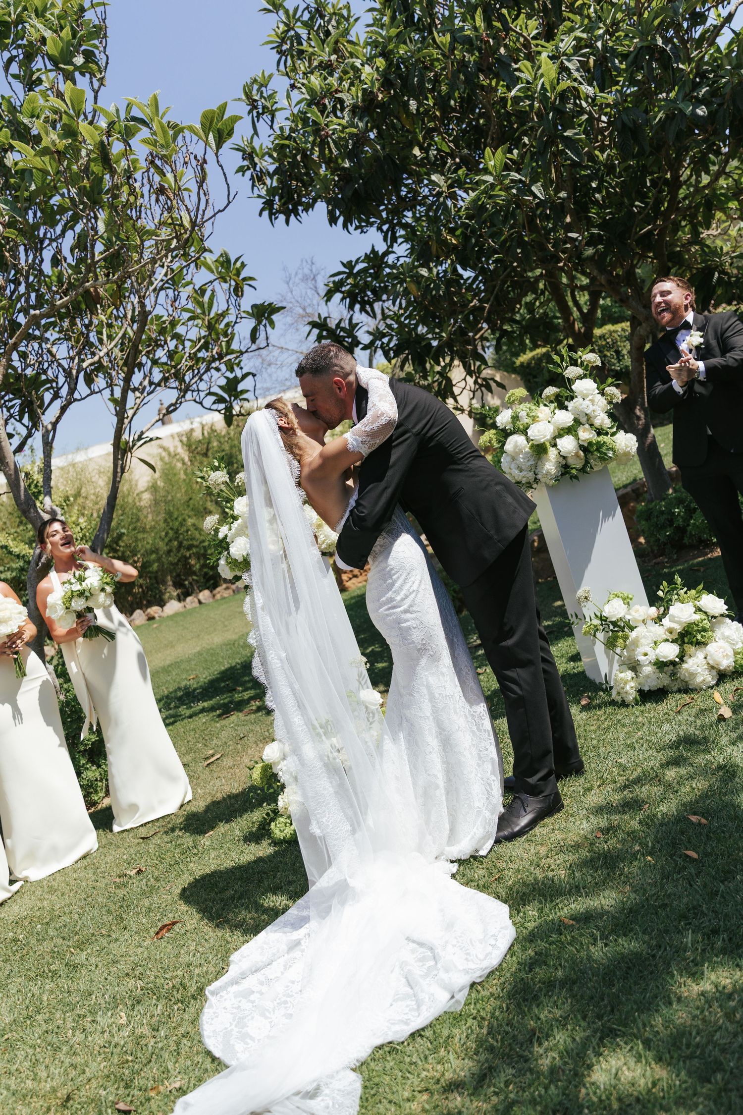 Bride and groom share a romantic first kiss at outdoor wedding ceremony with white floral arrangements and bridesmaids.