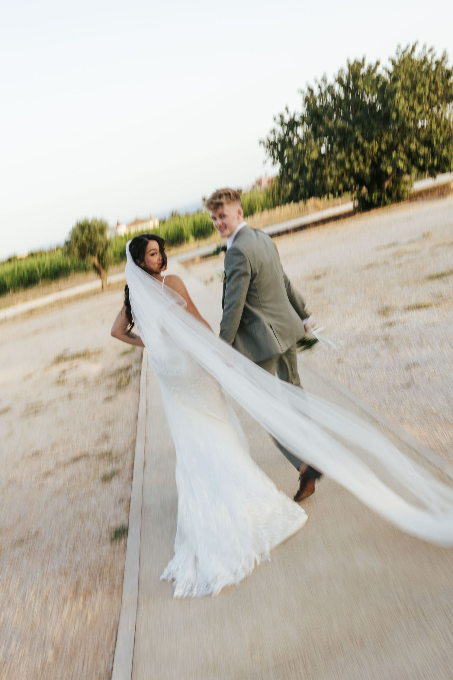 Bride in flowing white veil walks with groom in gray suit along outdoor path at golden hour wedding.