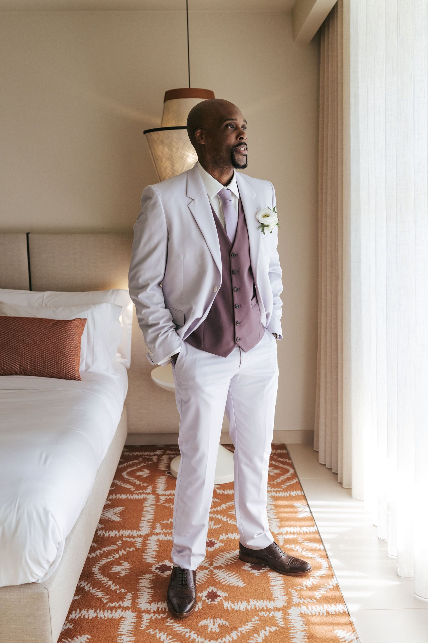 A person in a crisp white suit and mauve tie stands on a patterned orange and white rug in a minimalist room.