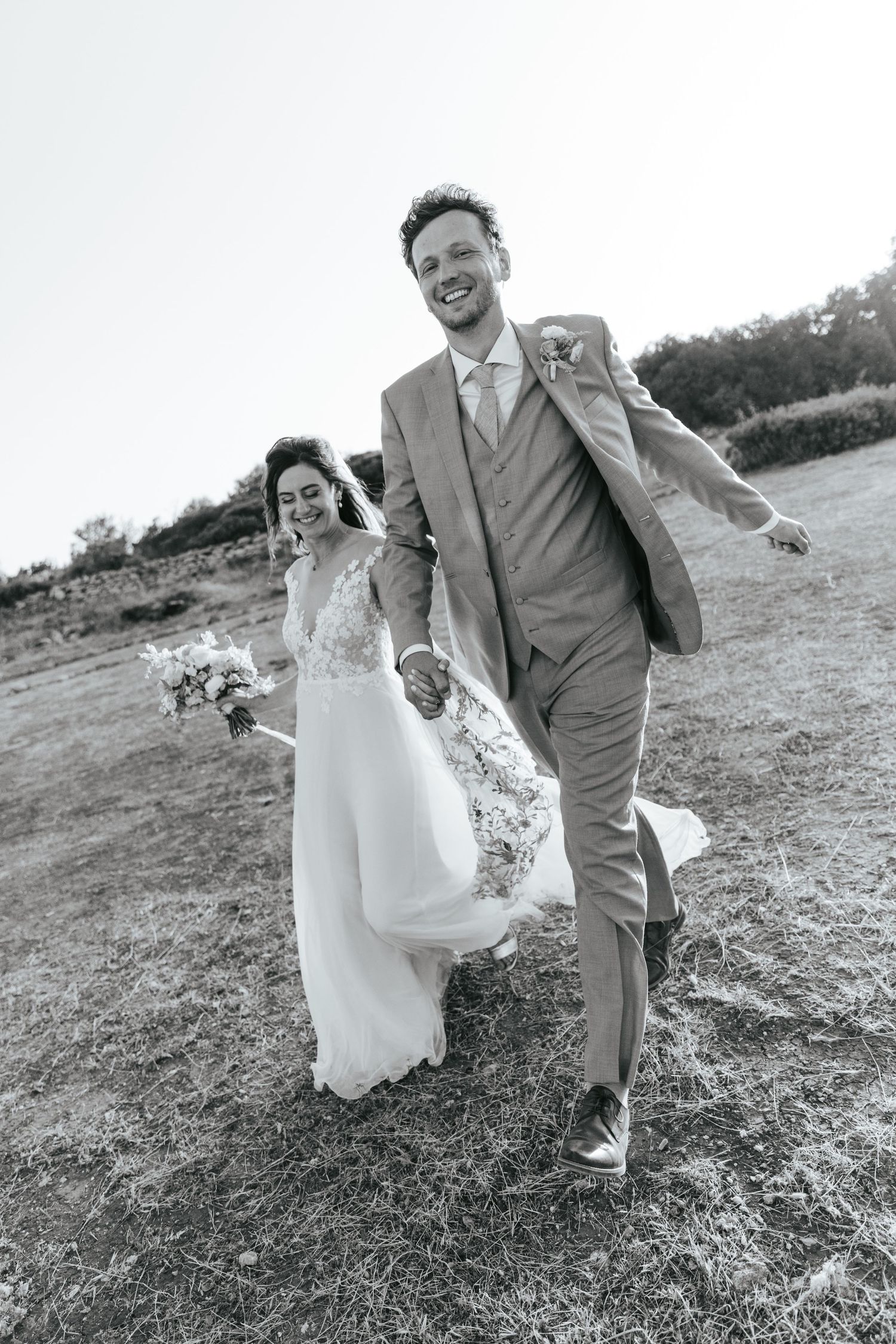 Black and white photo of a joyful bride and groom holding hands and walking through a field on their wedding day.