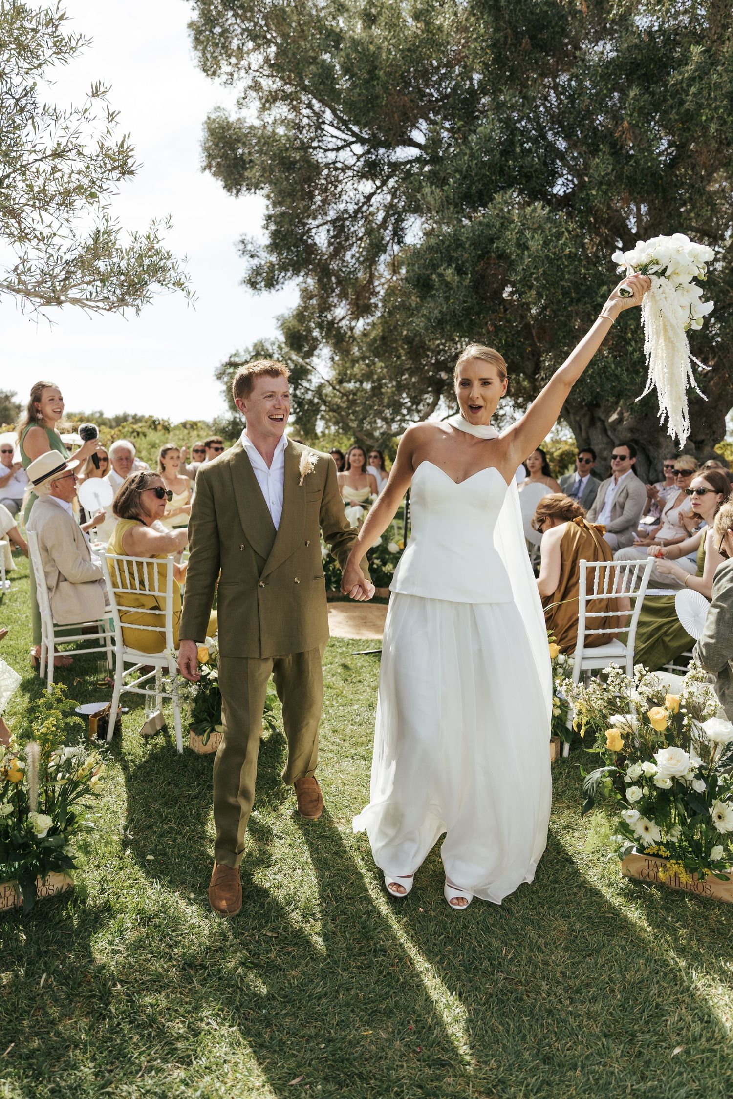 Joyful wedding couple celebrates with guests under olive trees during an outdoor garden ceremony.