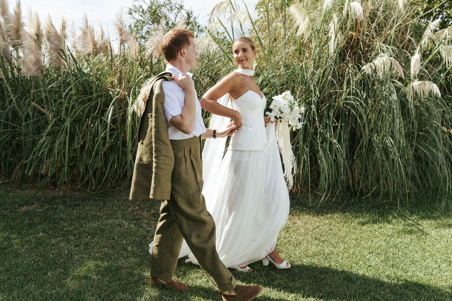 Bride in white gown and groom in olive suit walking together on green lawn with tall pampas grass backdrop.