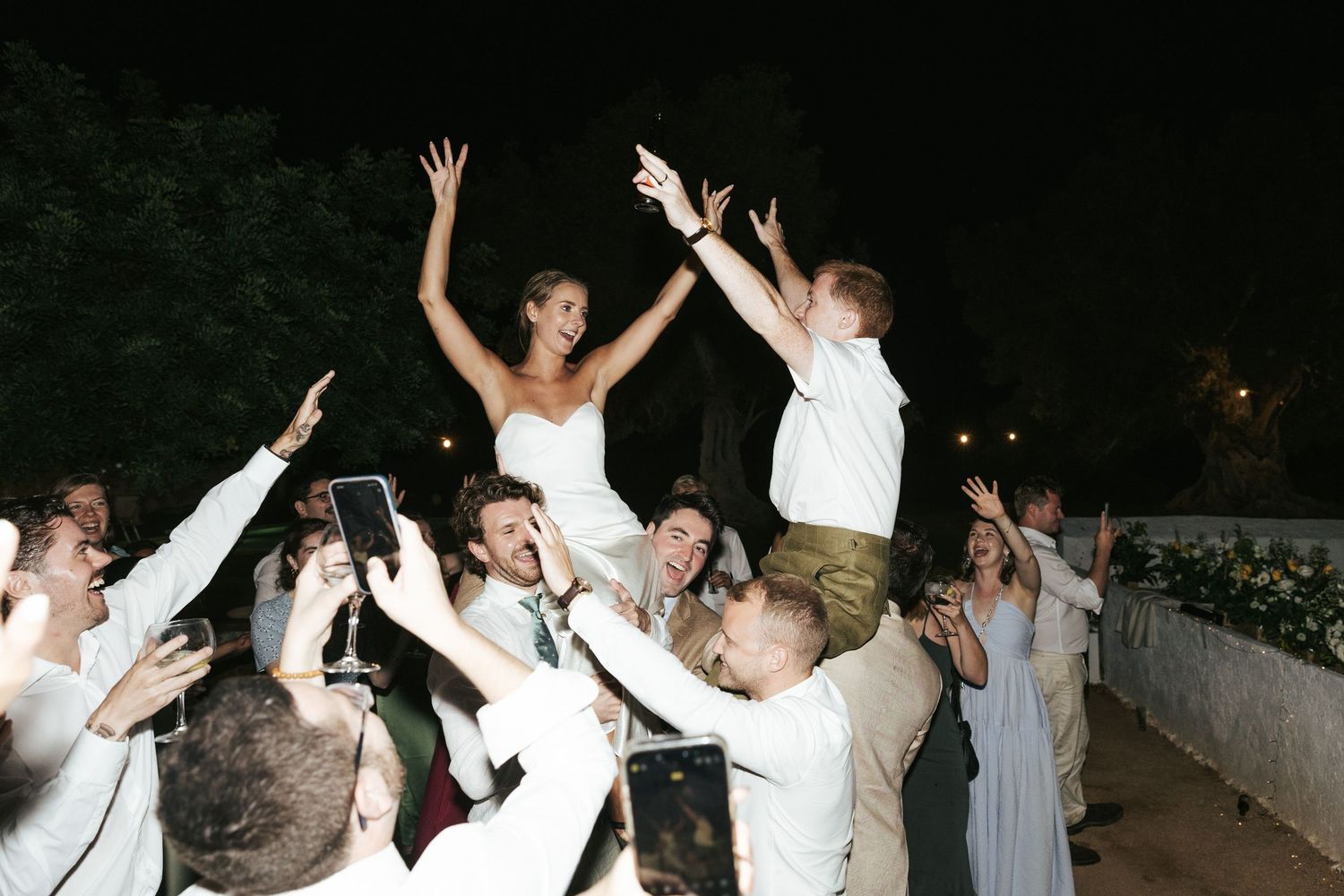 Bride and groom lifted by cheering wedding guests in a lively outdoor celebration.