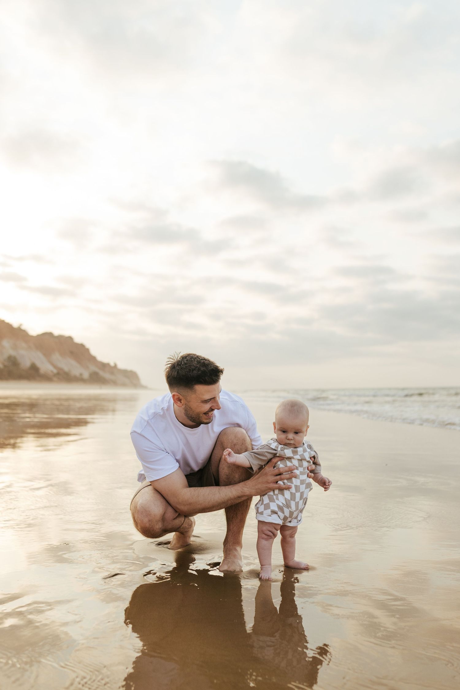 A parent crouches with a baby at the beach during golden hour sunset, with calm ocean waves and cliffs in background.