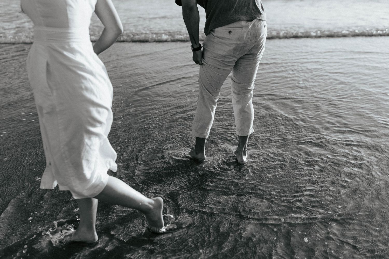 Black and white image of two people standing at the edge of water with their rolled up pant legs getting wet.