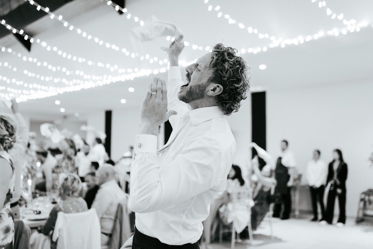 A guest in a white dress shirt raises a glass in celebration at an indoor wedding reception with string lights above.
