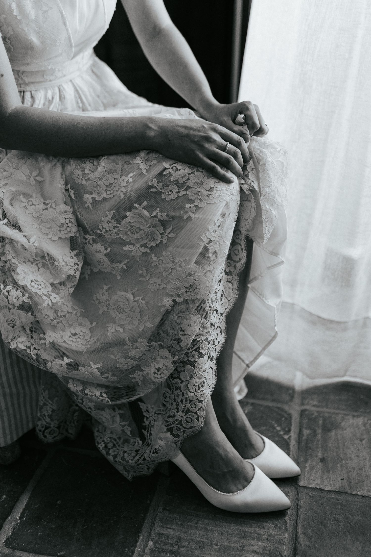 Black and white photo showing person in lace wedding dress adjusting white heels while sitting on dark floor.