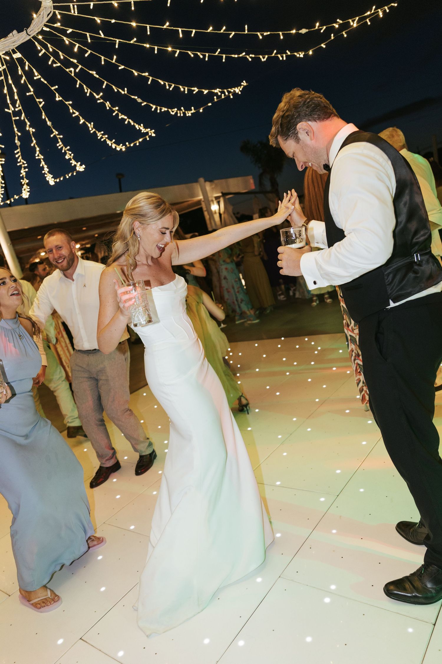 Bride and groom dancing joyfully on a glowing white dance floor under fairy lights at an elegant evening wedding reception.