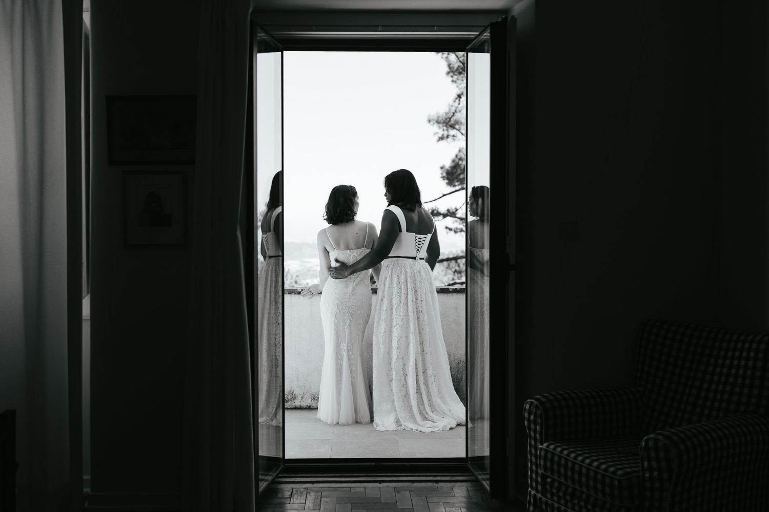 Two brides in white gowns stand together on a sunlit balcony, captured in elegant black and white photography.