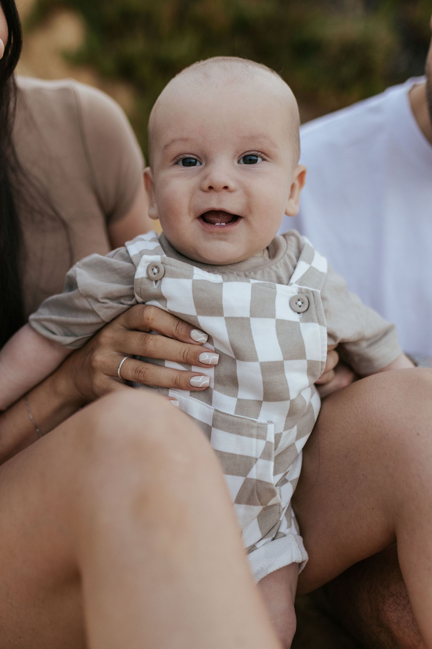 A smiling baby wearing a checkered beige and white outfit is held in someone's lap during an outdoor photo session.