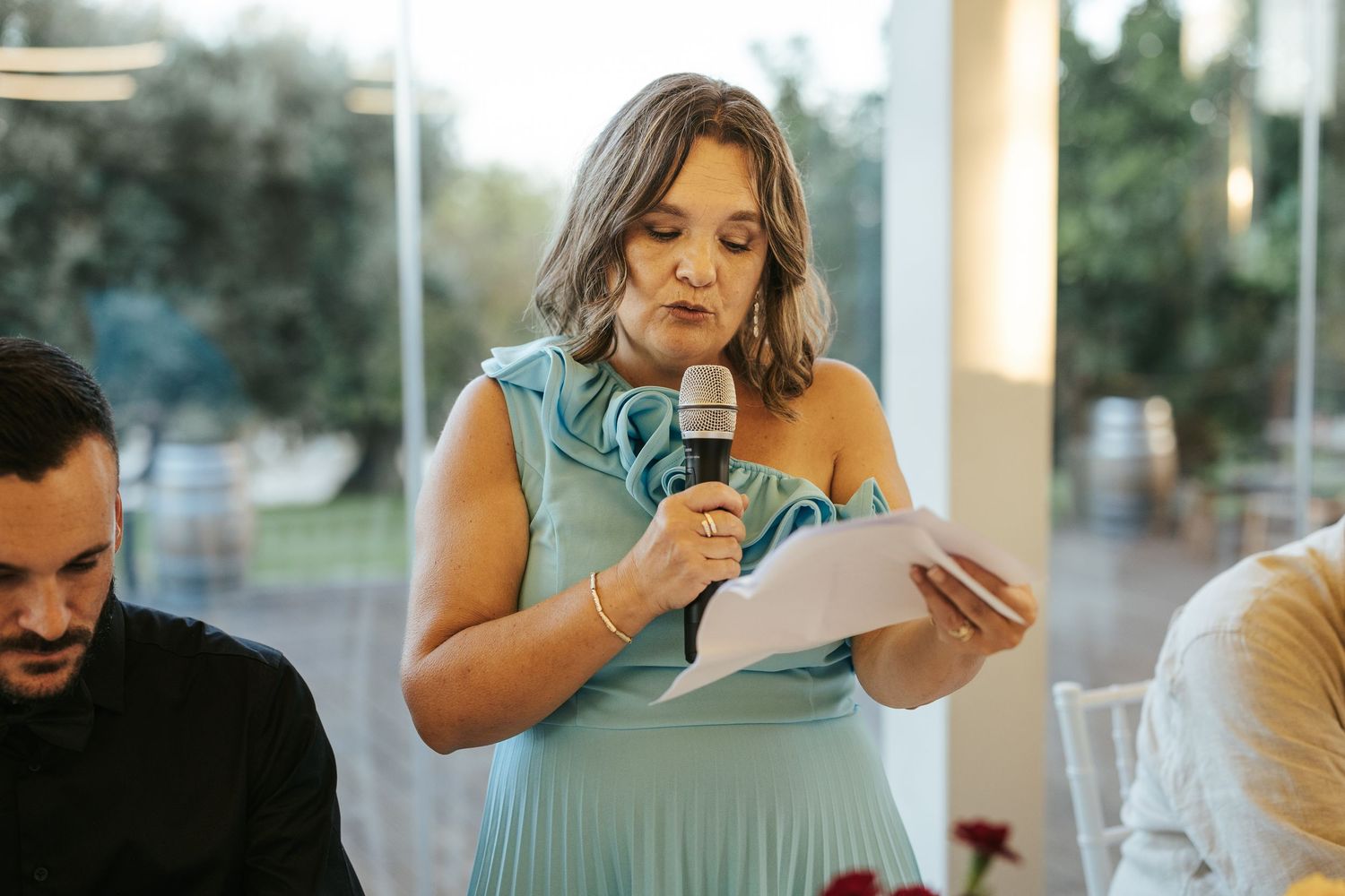 A person in a light blue dress holds a microphone while reading from papers at an indoor event.